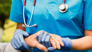 Standing nurse in blue uniform, wearing stethoscope and latex gloves hold hand of seated older patient.