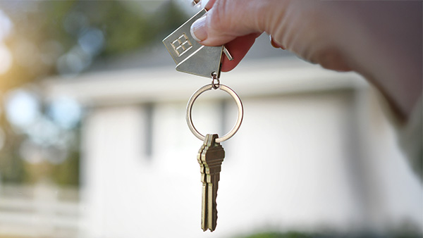 Hand dangling house keys with white home in background.