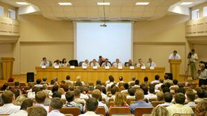 Government meeting room with citizens seated in foreground and officials on raised dais in background.